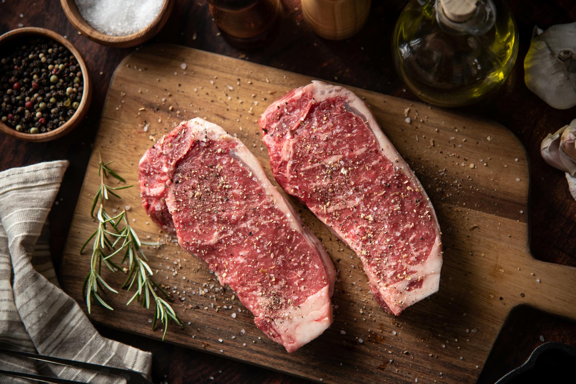Two seasoned raw strip steaks on a wooden cutting board